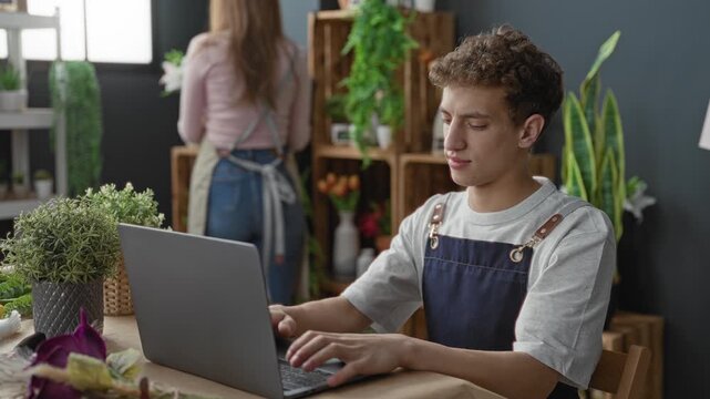 Man florist in apron types on a laptop in a flower studio while woman arranges plants on wooden shelves; small business productivity.