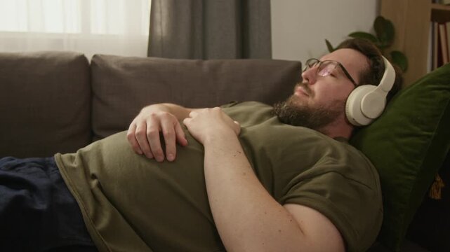 Tilt up shot of Caucasian adult man with diabetes lying on couch, wearing headphones and listening to music while wearing CGM sensor. Ideal for modern healthcare and diabetes management commercials