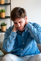 Fototapeta premium Man in denim shirt sitting on a sofa holding his neck in pain, showing discomfort and stress at home, portrait expressing neck pain, headache, tension, and seeking relief or medical attention.