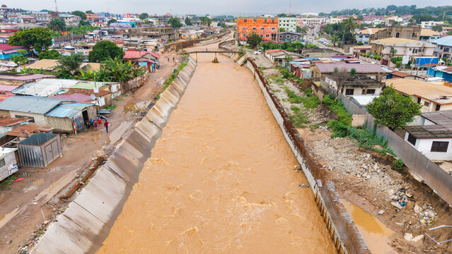 Aerial view, Muddy floodwater rushes through a concrete storm drainage channel in a densely populated residential neighborhood in Accra, Ghana