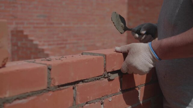 Close-up of a construction worker gloved hands, laying red bricks with wet cement. Professional bricklayer at the construction site.