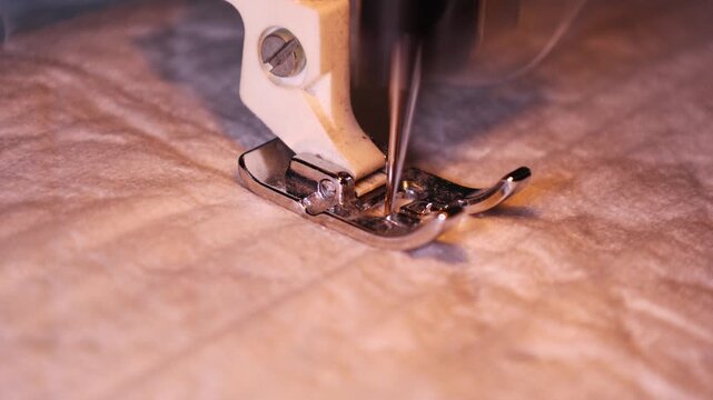 Detailed close-up of a sewing machines presser foot and needle sewing fabric. The metal components are focused, capturing the stitching process on a piece of soft, light-colored cloth under warm.