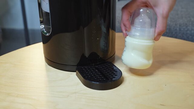 Close up shot of a hand holding a plastic baby bottle filled with milk next to a modern black formula maker machine. The items sit on a light wooden table in a domestic indoor home setting.