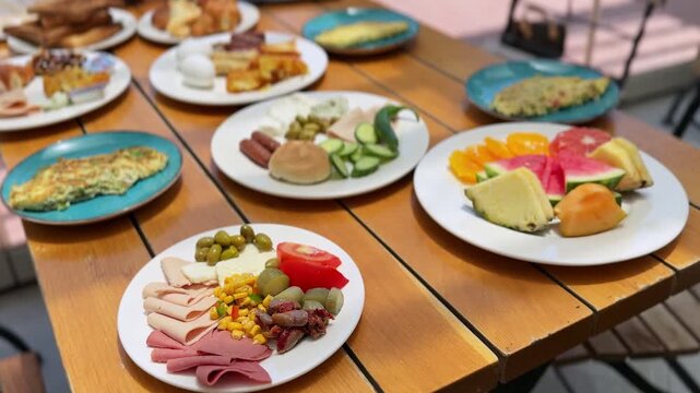 Static shot of a large breakfast buffet spread on an outdoor wooden table. Plates of fresh fruit, omelets, pastries, and cold cuts sit in bright morning daylight at a patio cafe.