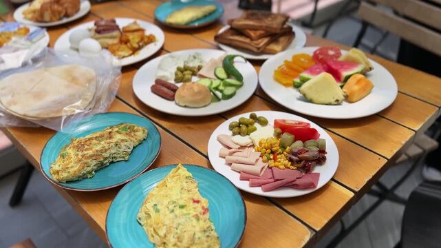 A hand places a white plate of cold cuts and olives onto a wooden table filled with a large breakfast buffet spread. Fresh fruit, omelets, and bread sit in bright morning daylight on a sunny patio.