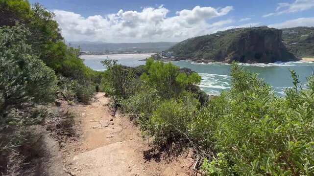 A narrow dirt path winds through lush greenery along the cliffs of Knysna, offering views of the lagoon and rocky coastline. South Africa.