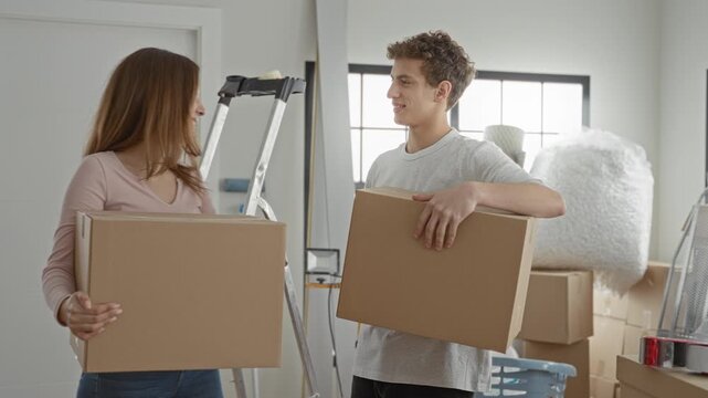 Couple holding moving boxes with arm around shoulder and smiling while lifting cartons inside a building; moving day joy together.