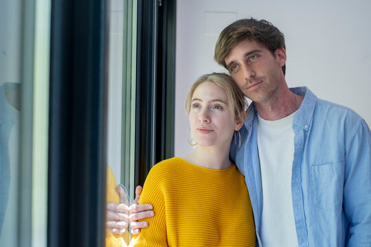 Young couple standing together by a window, looking thoughtfully outside in a cozy home, sharing a quiet affectionate moment of love and hope