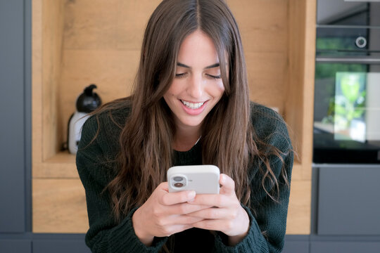 Smiling young woman in a cozy kitchen looking down at her smartphone, texting or browsing with a relaxed expression, long brown hair and casual sweater conveying modern home lifestyle and happiness