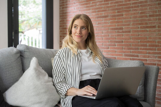 Young woman working on laptop at home on a gray couch by a brick wall, looking thoughtful and relaxed while using her computer
