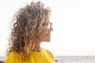 Profile of smiling woman with curly hair and glasses wearing a yellow shirt by the sea at sunset, thoughtful and relaxed seaside portrait © simona