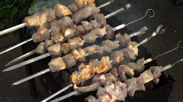 Pork shish kebab on metal skewers appetizingly roasting on a mangal grill, high angle view. The process of cooking marinated meat over hot wood charcoals with rising white smoke.