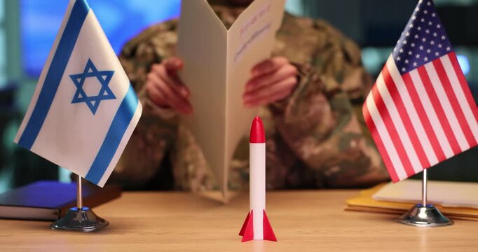 Military officer carefully studies secret strategic documents. Detailed toy rocket stands near flags of United States and Israel. Slow motion