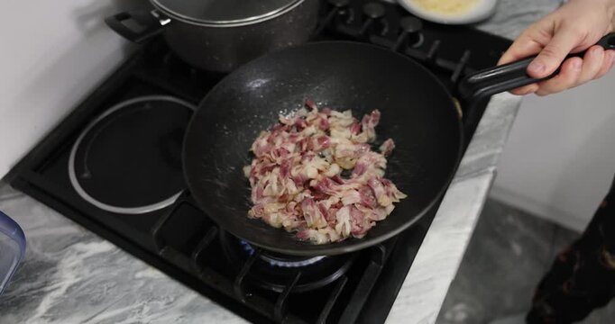 Experienced chef carefully stirs crispy bacon by shaking heavy frying pan. Juicy pork meat slowly becomes golden and crispy. Slow motion