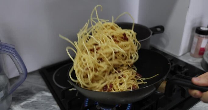 Chef mixes tender spaghetti with crispy bacon by shaking heavy black frying pan over stove. Aromatic steam rises through kitchen. Slow motion