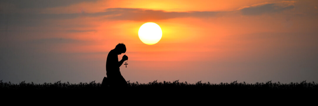 A silhouette of a man kneeling in prayer against a dramatic sunset, with the sun casting a warm glow over the landscape