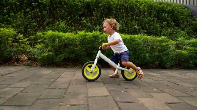 Playful toddler rides balance bike on sidewalk in slow motion side-tracking shot. Little puppy joyfully runs ahead, young child accelerate to catch up canine companion.