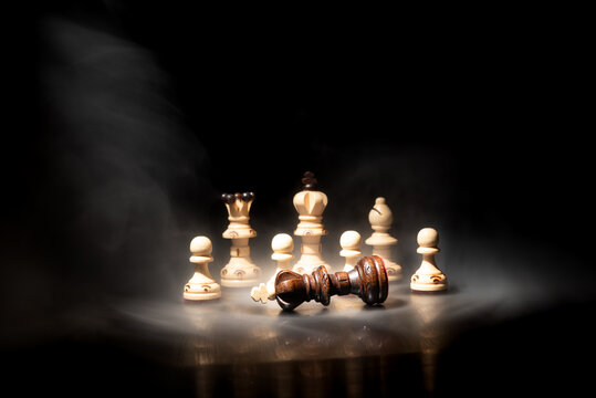 Dark chess king fallen among white pieces in dramatic smoke and spotlight. Concept of checkmate, defeat, leadership, business strategy and competition.