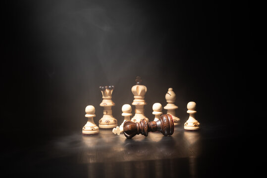 Dark chess king fallen among white pieces in dramatic smoke and spotlight. Concept of checkmate, defeat, leadership, business strategy and competition.