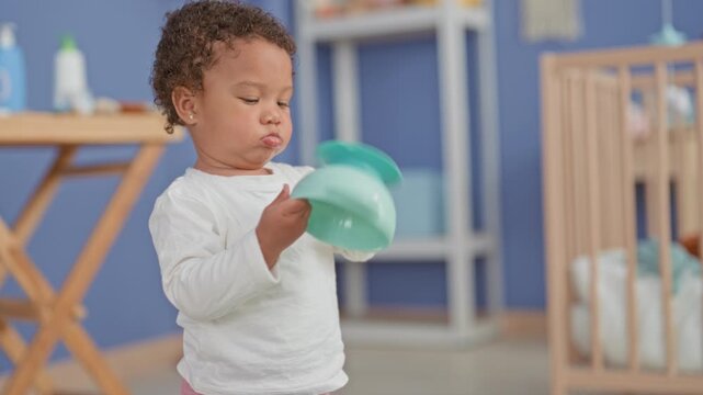 Little girl holding teal bowl and touching its rim while sitting in a nursery inside a building; quiet curiosity.