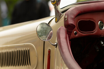 Naklejka premium Close-up view of a cream-colored vintage car showing the dashboard, burgundy leather steering wheel, round side mirror, vented door panel and chrome trim