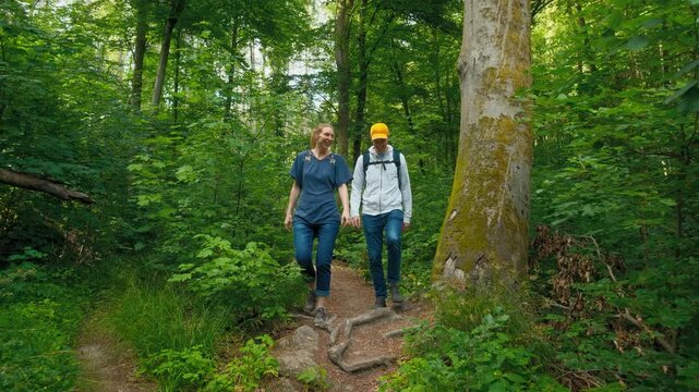 Two happy adults hiking, talking and laughing on a narrow scenic path in a lush green forest, slow motion