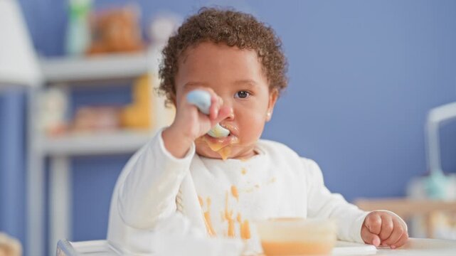 African american little girl eating with a spoon in a building highchair, bib and mouth messy with orange puree and blue wall behind; curiosity independence.