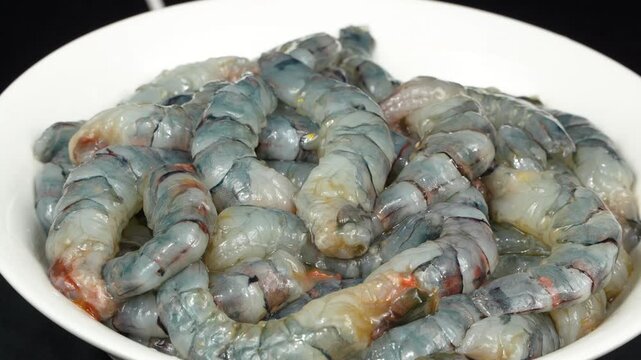 A horizontal view of freshly peeled and headless black tiger shrimp piled on a white plate, slowly rotating.