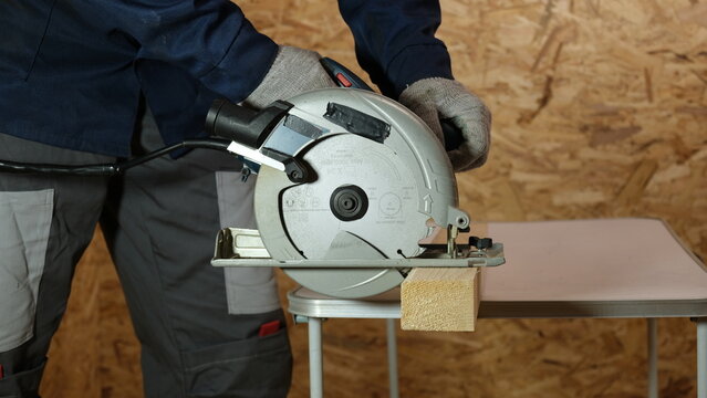 Builder saws a board with a circular saw cutting a wooden plank in a joinery. Carpenter uses portable circular saw tool to cut wood at wooden table in workshop. Worker sawing pine board. Real image