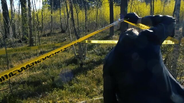 Male police detective wearing a black leather jacket and gloves crossing a yellow tape to enter an outdoor crime scene in a sunlit forest, carefully inspecting the area for evidence