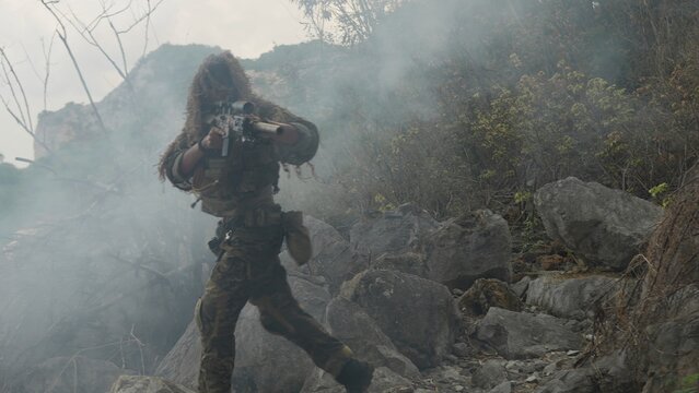 A special forces soldier in a ghillie suit advances through a dense smoke screen on a rocky mountain, conducting a covert tactical military operation.