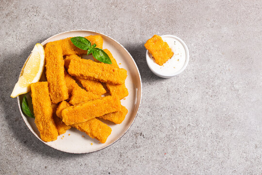 Tasty golden-brown fish fingers served with dip and lemon slices. Crispy breaded fish sticks on light background. 