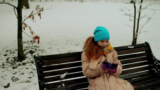 little beautiful girl in a winter jacket sits on a bench and dials a number on the phone in a snowy city park