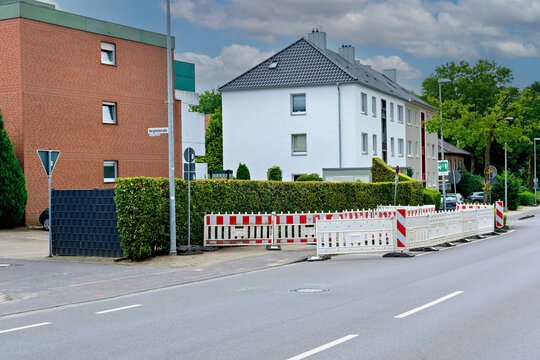 A street scene featuring modern residential buildings, green hedges, and temporary plastic safety barriers for roadworks or construction under a cloudy sky.