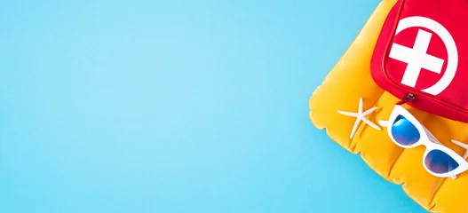 Summer flat lay with red medical first aid kit bag, sunglasses, and starfish on yellow inflatable mattress on blue background. Safe beach travel vacation concept with broad empty copy space. © prime1001