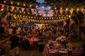 Obraz premium Outdoor neighborhood block party at night with people dining under string lights and flag buntings.