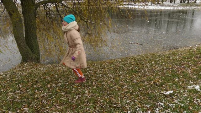 Small joyful beautiful girl runs near frozen pond in old city park.