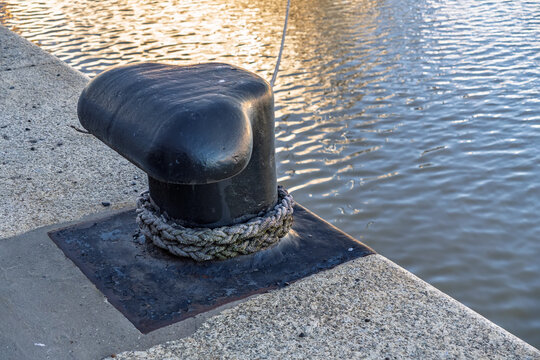 Heavy-duty black metal mooring bollard on a concrete pier with a thick rope, overlooking calm river water with golden reflections during sunset