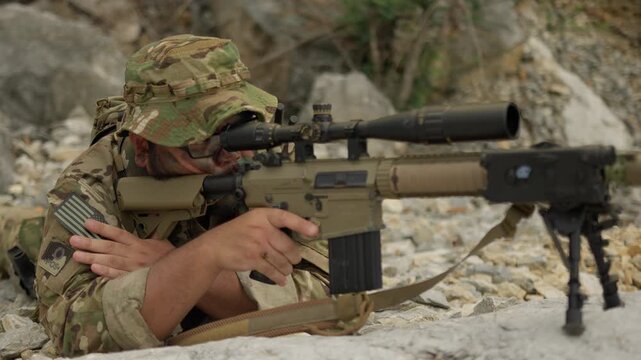 A military sniper in multicam camouflage aims his rifle from a concealed rocky position, conducting surveillance during a special operations mission.
