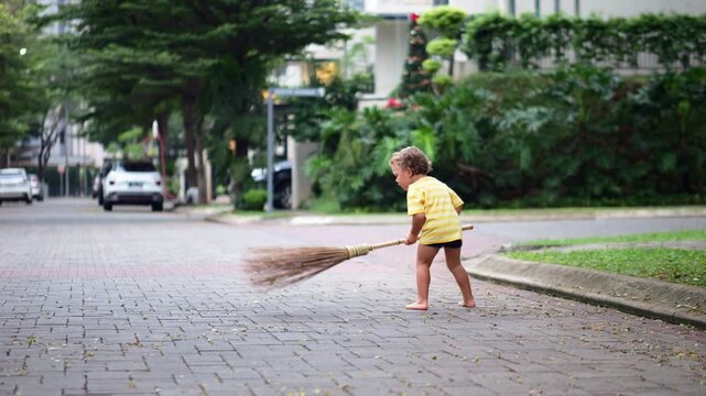 Young child diligently sweeps road from small leaves with big broom, energetically imitating adults and making large swings. In midst of this process, playful puppy becomes adorable hindrance