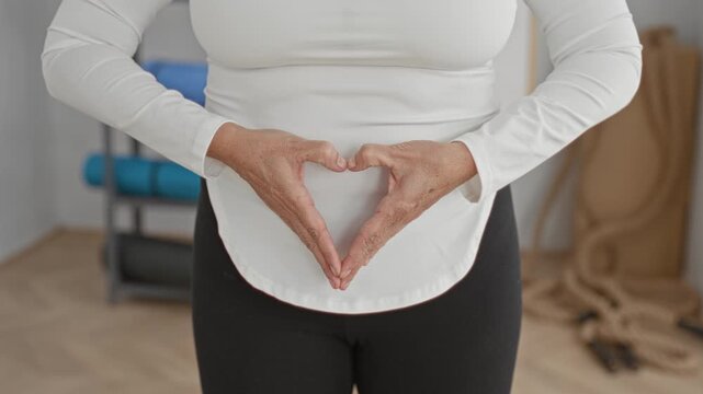 Middle age hispanic woman forming heart with hands over abdomen wearing white long sleeve top and black leggings in yoga studio; serenity wellness prenatal.