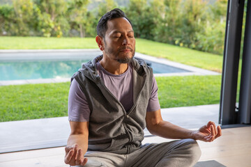 Fototapeta premium Mature Asian man meditating on yoga mat in activewear by sliding wall overlooking pool and lawn