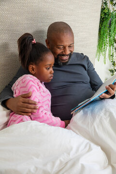 African American father dark shirt and daughter pink chevron robe sitting on bed reading storybook