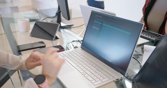 Diverse female coworkers leaning at desk spotting code, pointing, typing on silver laptop, pausing
