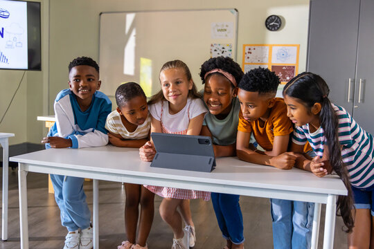 Diverse school-age children leaning over tablet on white table in classroom, wearing casual clothes