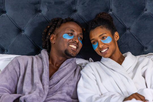 African American couple reclining on bed with tufted headboard, wearing robes and blue gel patches