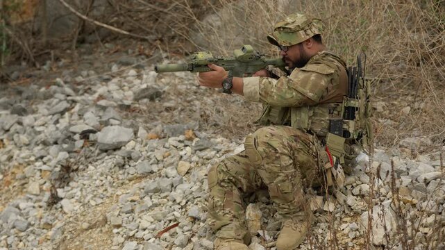 A fully equipped special forces soldier takes a tactical position, aiming his assault rifle during a reconnaissance mission in a rugged, arid environment.