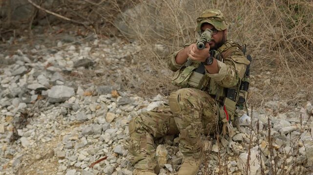 A special forces soldier aims his assault rifle from a defensive position on a rocky hillside, conducting surveillance during a military combat operation.