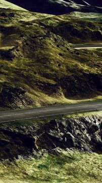 A road is visible cutting through green hills. The landscape features rocky terrain and patches of grass. The scene shows clear weather and bright sunlight.