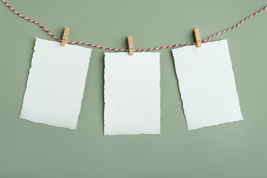 Close-up shot of white blank sheets of watercolor paper with torn edges suspended on decorative rope using small wooden clothespins on a green board.